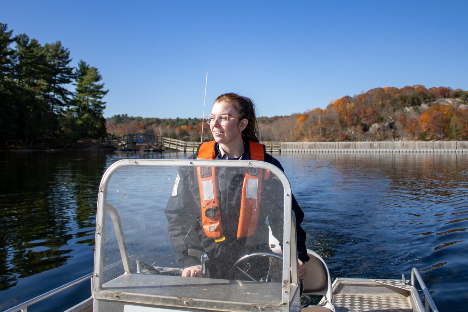 A person wearing a PFD driving a small motorboat in a lake, with a fall-coloured forest behind them.