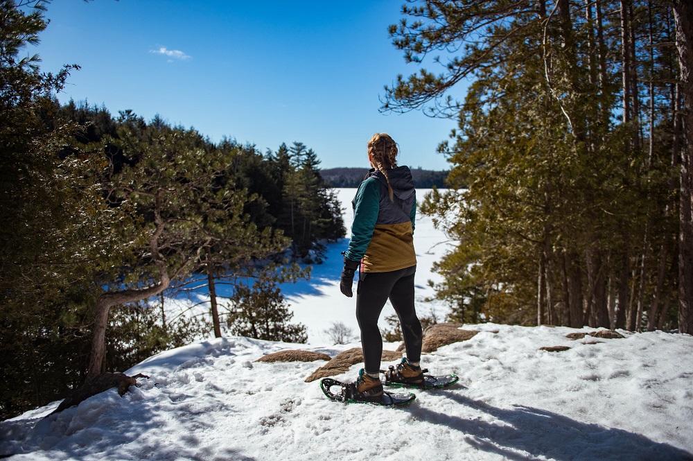 A person wearing snowshoes on a snow-covered lookout in a forest, looking over a frozen lake on a sunny day