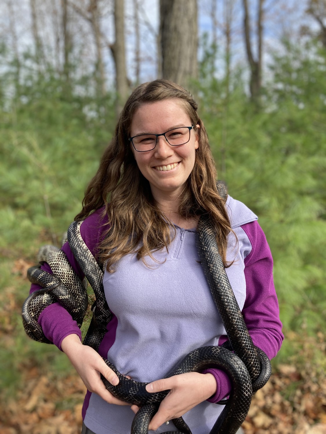 A person in a purple jacket with a very long grey snake coiled around their right arm, up around their neck, and down to their left arm. They are smiling at the camera.