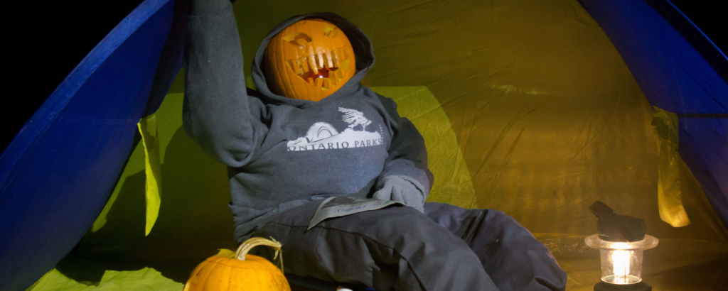 A campsite decorated for Halloween that shows a pumpkin-headed mannequin dressed up as a camper sitting in front of a tent with another pumpkin beside them.