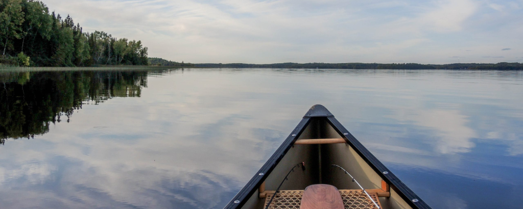 The bow of a canoe with a paddle resting across the empty seat, on a lake where the evening sky reflects in the water