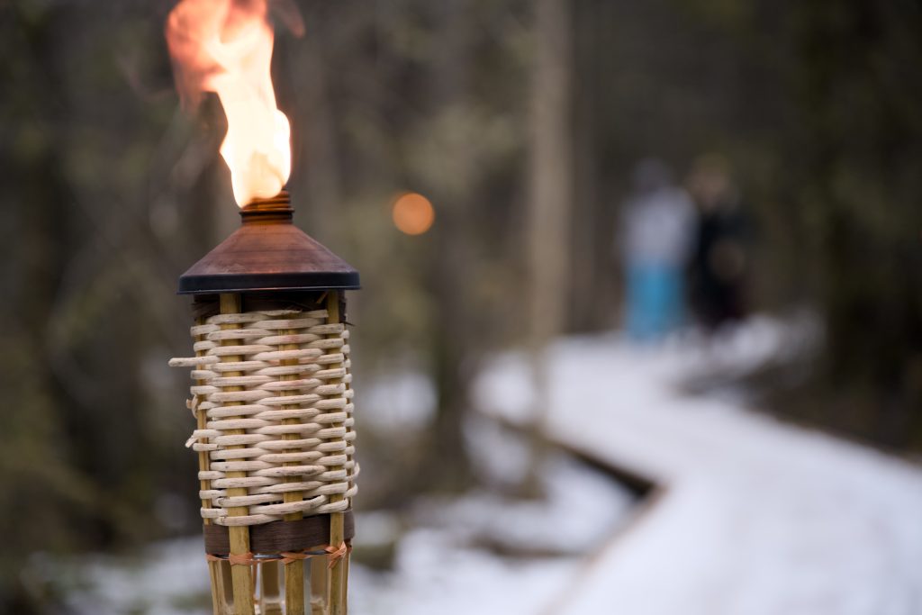 A flaming torch, with two people walking on a snowy boardwalk trail through the forest in the background