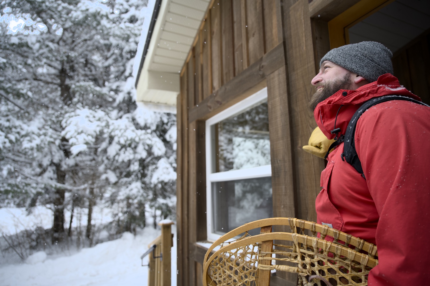 Un homme sortant d’une cabane et tenant des raquettes en se dirigeant vers une forêt hivernale