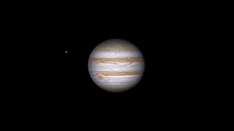 Jupiter in the black of space. It looks like a giant orange and white striped sphere, with the red dot on its lower left side visible