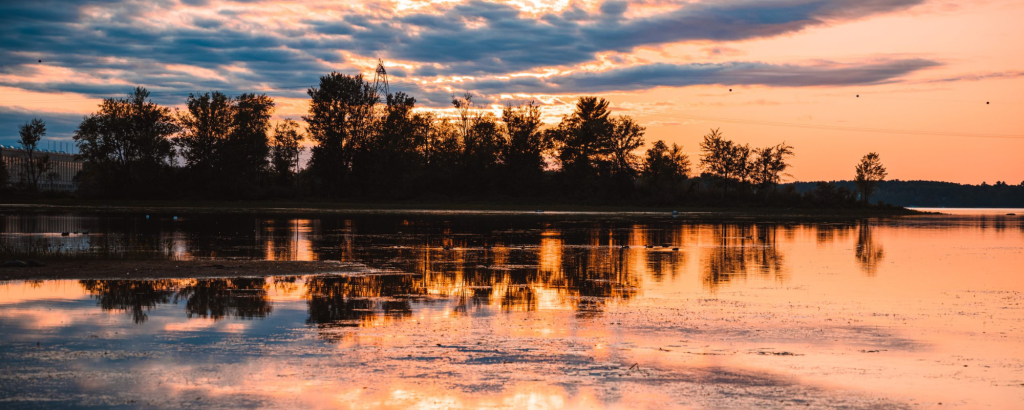 An orange sunset reflecting off a lake with a forested shoreline in the distance