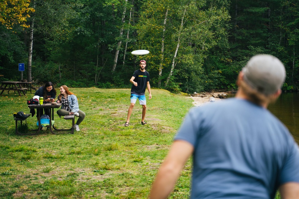 two people playing frisbee, people sitting on picnic table in background