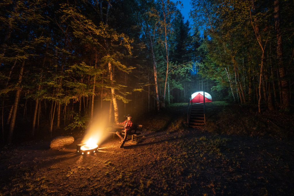 person sitting on bench around campfire at night. Lit tent in forest in background