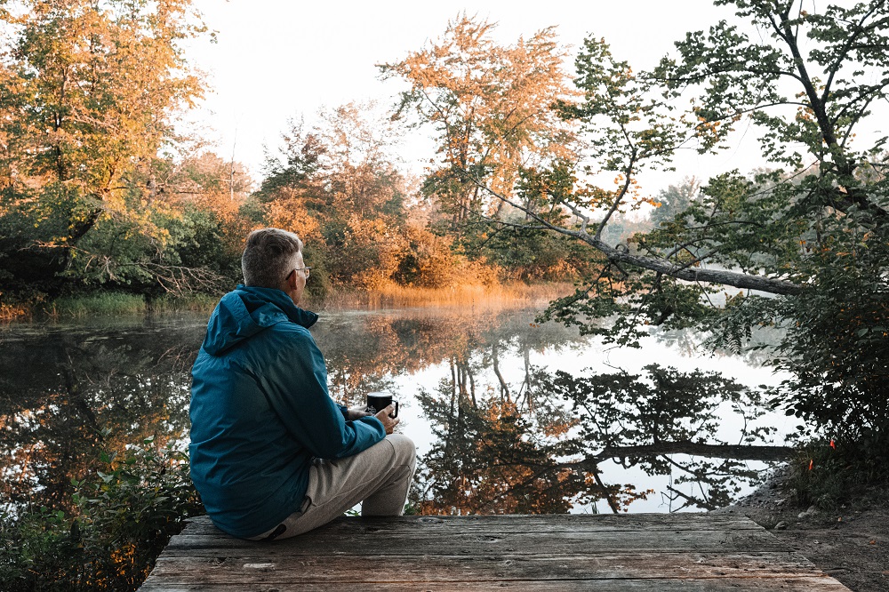 person sitting on edge of river drinking coffee, looking out at lake