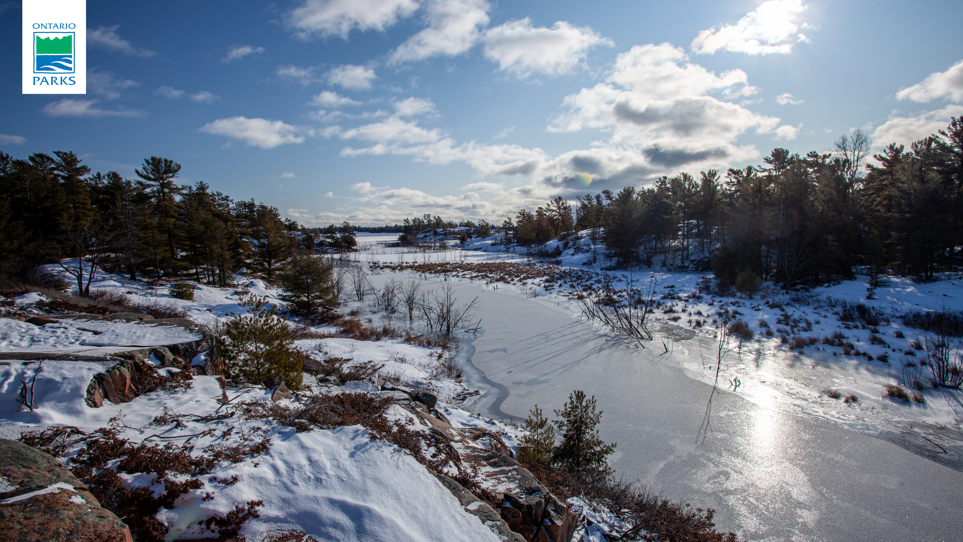 A frozen river between two rocky and lightly forested shorelines covered in snow on a sunny day