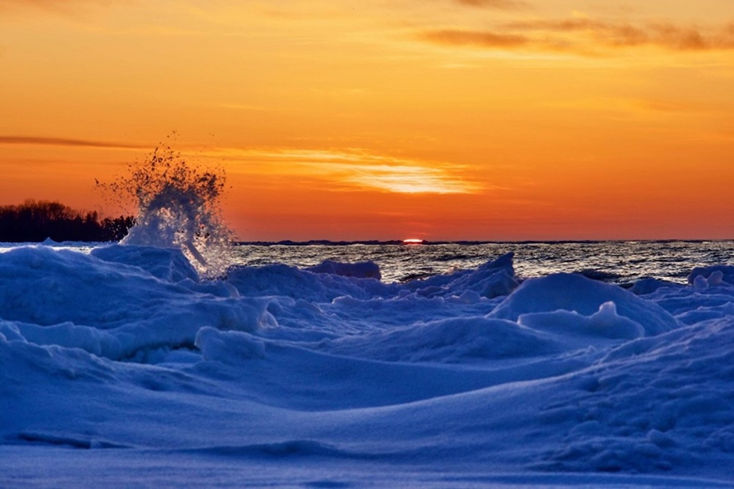 A vivid orange sunset on a lake with ice and splashing water in the foreground. The ice appears blue.