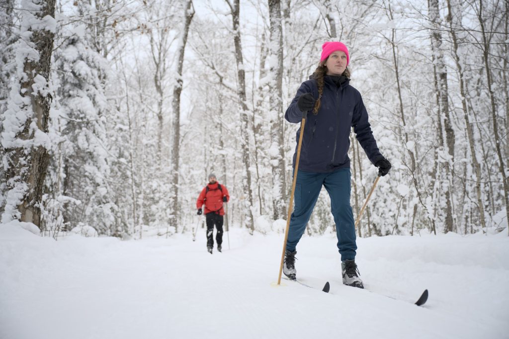 woman cross-country skiing through snowy forest, another skier following behind her