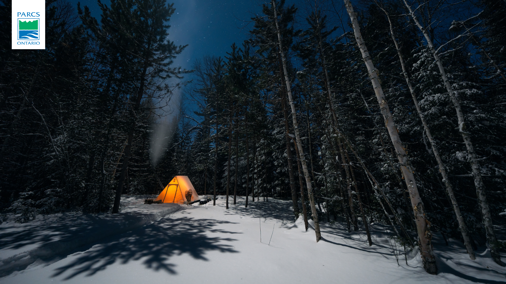 Une tente chauffée qui brille d’une lumière orange dans une forêt enneigée par une nuit d’hiver étoilée et éclairée par la lune. Le logo de Parcs Ontario est affiché dans le coin supérieur gauche de l’image.