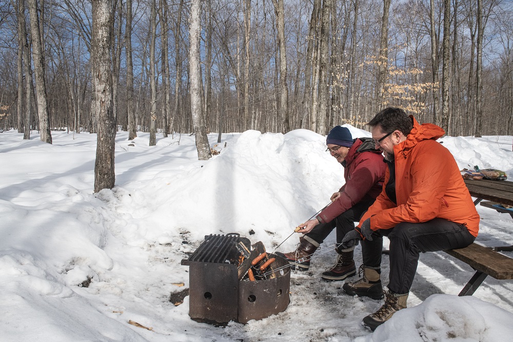 two people sitting on picnic table, roasting hot dogs over the fire