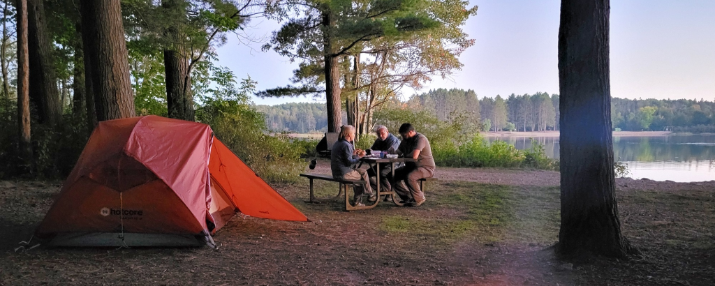 campers sitting at a picnic table beside a tent with a lake in the distance