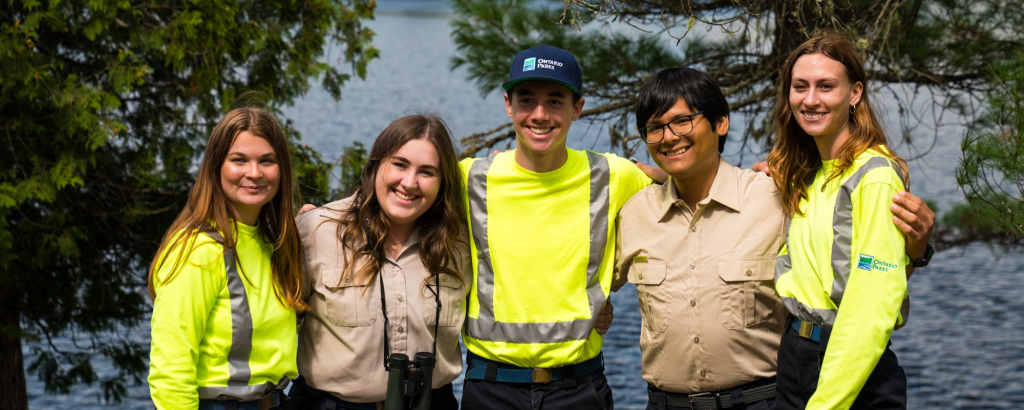 A group of smiling summer students, some wearing office uniforms, some wearing maintenance uniforms, standing on a beach in front of a river, their arms over each other's shoulders