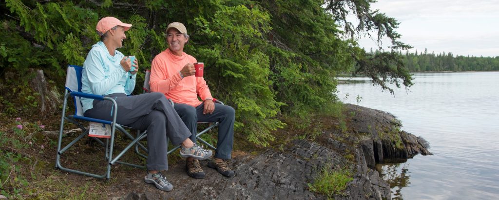 two people sitting on camp chairs at lake's shore, holding coffee