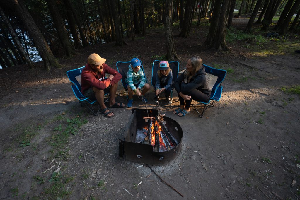 Une famille assise autour d’un feu de camp sur un emplacement de camping arboré.