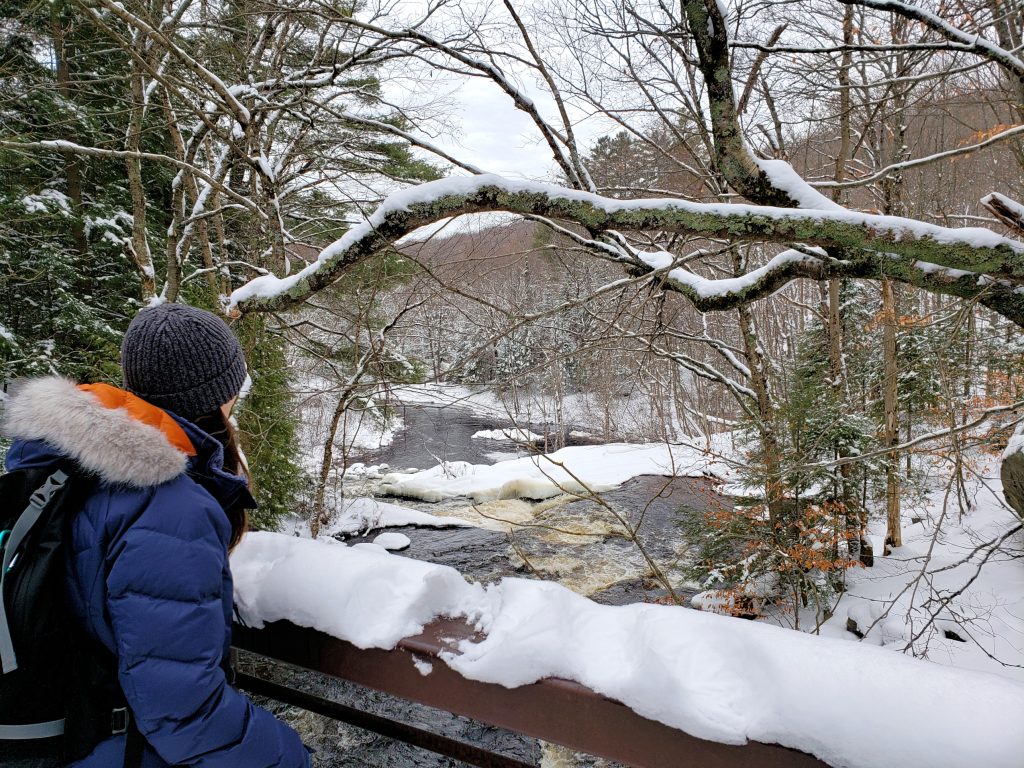 hiker looking over boardwalk at snow-covered forest with partially frozen river