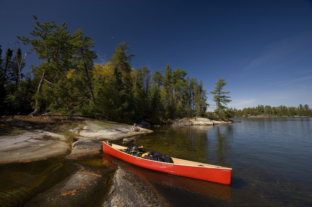canoe on rocky shore with forest, person sitting on shore looking at lake