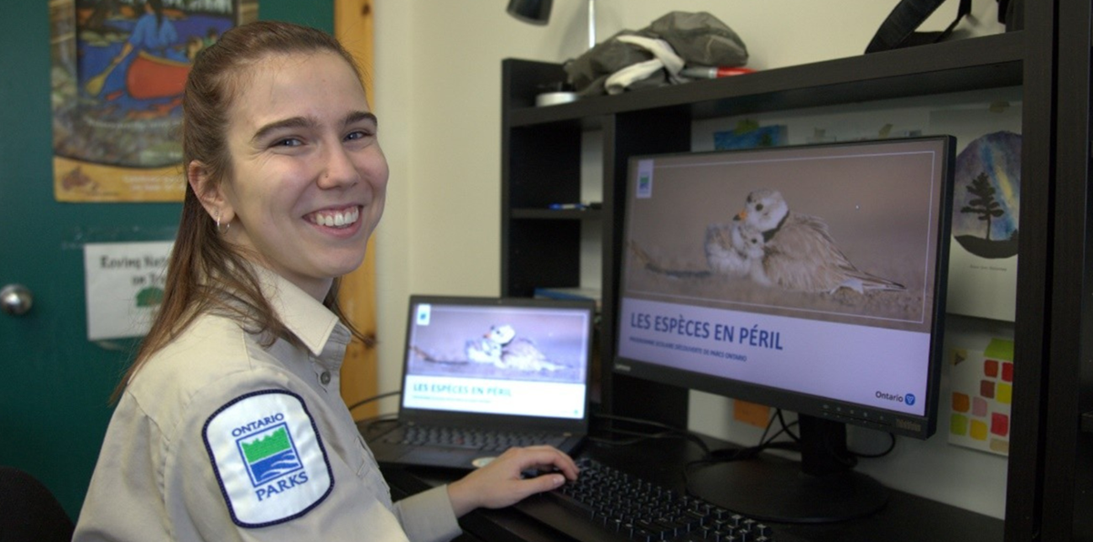 Marianne assise à un bureau devant deux ordinateurs, dont les écrans affichent une diapositive du programme virtuel pour la 4ème année: “Les espèces en péril”