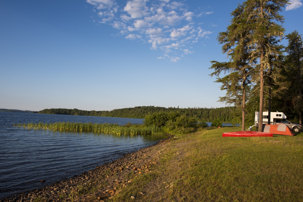 Un terrain de camping au bord d’un lac avec un VR, une tente et un canot
