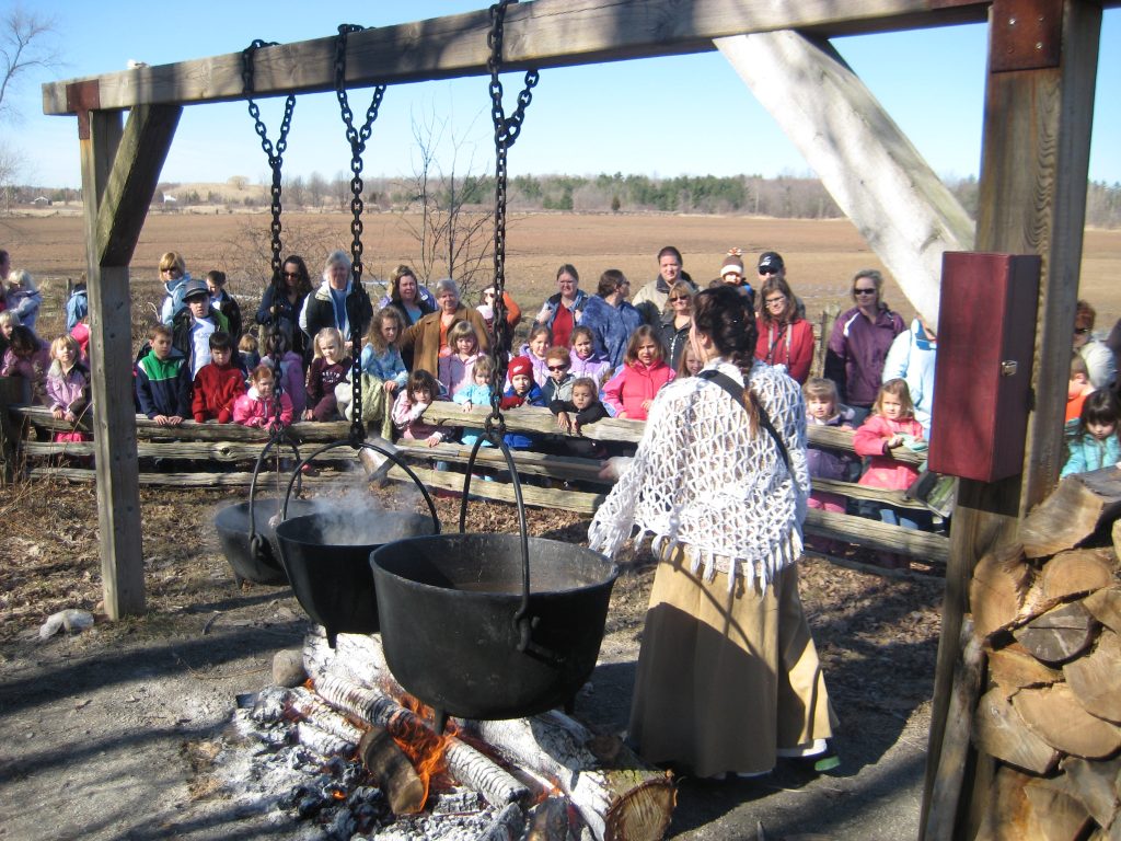 interpreter presenting in front of boiling sap in large cast iron pots. Large group of visitors watching