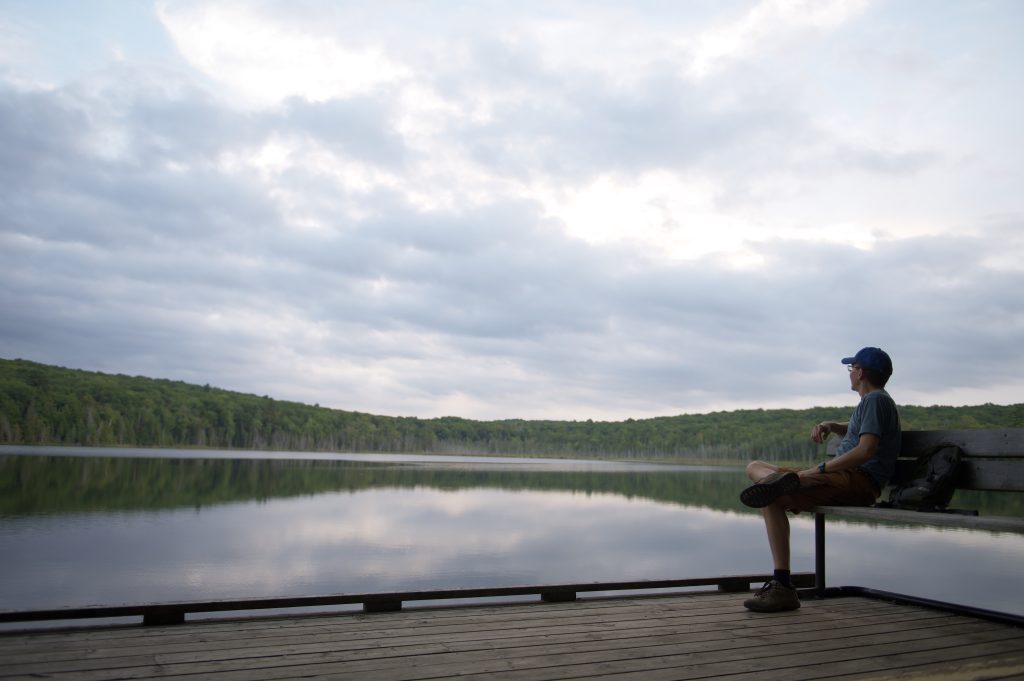 person sitting on bench, looking out at still lake with forested shore reflected