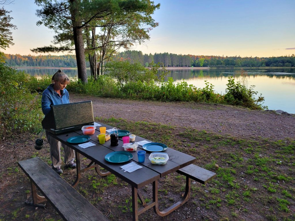 woman cooking at camp stove on picnic table on rocky day use area beside lake. Table is set for dinner.