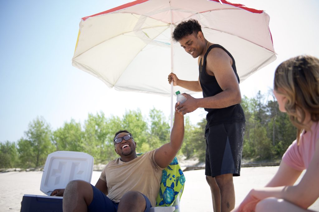man sitting in beach chair offering bottle to man holding umbrella. Woman watching in foreground