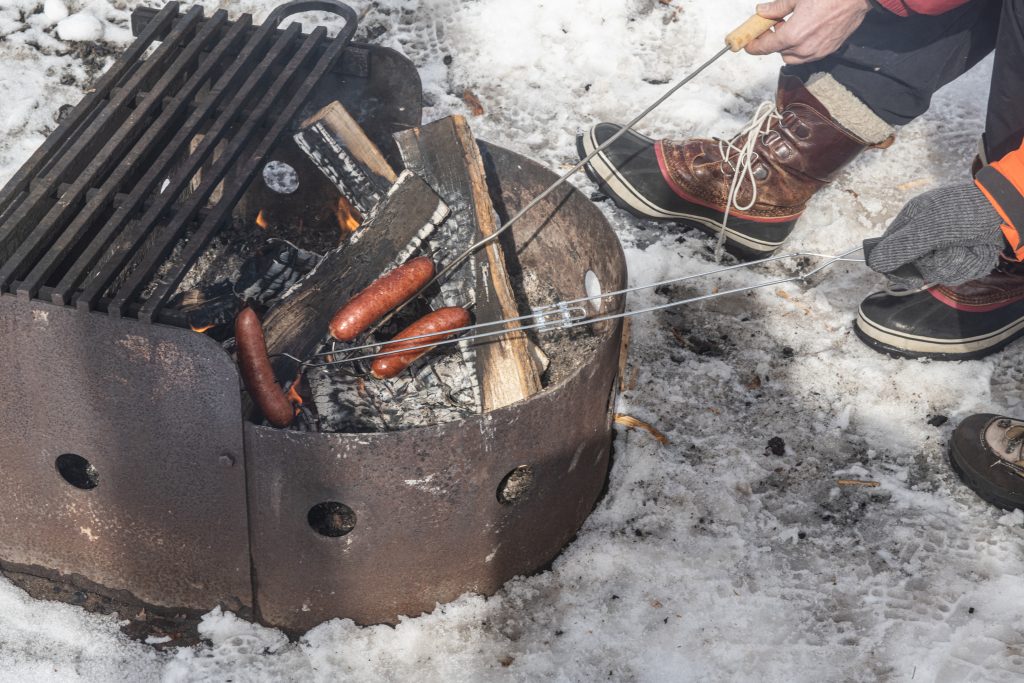 hands roasting hot dogs over campfire pit surrounded by snow